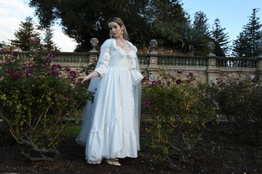 Tarihi beyaz gelinlik ve fantezi pelerin giyen güzel sarışın mankenin portresi. Peri masalı şatosu bahçesinin romantik gül bahçesinde gezinen ruhani figür.