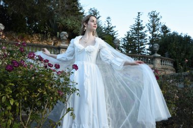 Tarihi beyaz gelinlik ve fantezi pelerin giyen güzel sarışın mankenin portresi. Peri masalı şatosu bahçesinin romantik gül bahçesinde gezinen ruhani figür.