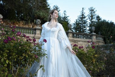 Tarihi beyaz gelinlik ve fantezi pelerin giyen güzel sarışın mankenin portresi. Peri masalı şatosu bahçesinin romantik gül bahçesinde gezinen ruhani figür.