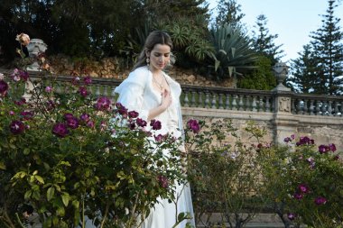Tarihi beyaz gelinlik ve fantezi pelerin giyen güzel sarışın mankenin portresi. Peri masalı şatosu bahçesinin romantik gül bahçesinde gezinen ruhani figür.