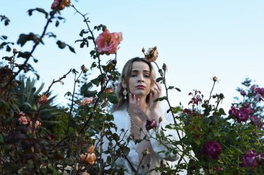 Beyaz gelinlik giymiş, elleri bükülmüş, dikenli gül çalılarının arasından kameraya doğru uzanan, jestsel pozlar veren, mavi gökyüzü arkaplanlı güzel sarışın mankenin portresi..