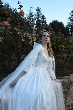 Tarihi beyaz gelinlik ve fantezi pelerin giyen güzel sarışın mankenin portresi. Peri masalı şatosu bahçesinin romantik gül bahçesinde gezinen ruhani figür.