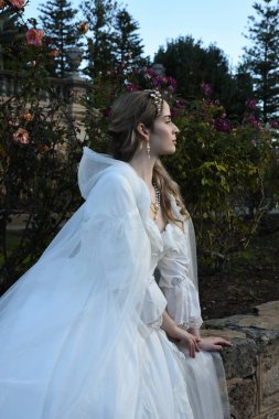 Tarihi beyaz gelinlik ve fantezi pelerin giyen güzel sarışın mankenin portresi. Peri masalı şatosu bahçesinin romantik gül bahçesinde gezinen ruhani figür.