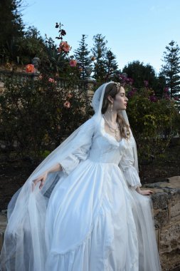 Tarihi beyaz gelinlik ve fantezi pelerin giyen güzel sarışın mankenin portresi. Peri masalı şatosu bahçesinin romantik gül bahçesinde gezinen ruhani figür.