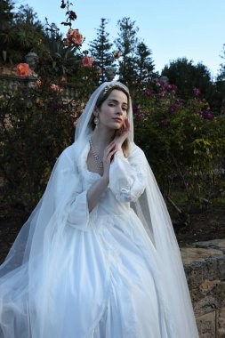 Tarihi beyaz gelinlik ve fantezi pelerin giyen güzel sarışın mankenin portresi. Peri masalı şatosu bahçesinin romantik gül bahçesinde gezinen ruhani figür.