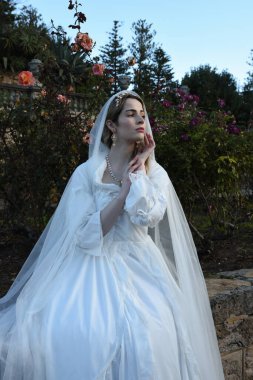 Tarihi beyaz gelinlik ve fantezi pelerin giyen güzel sarışın mankenin portresi. Peri masalı şatosu bahçesinin romantik gül bahçesinde gezinen ruhani figür.