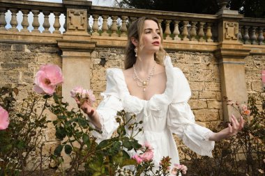 Güzel sarışın mankenin portresi tarihi fantezi beyaz gelinlik giymiş, elleri güllere uzanan, peri masalı kale bahçesindeki gül bahçesi arka planında