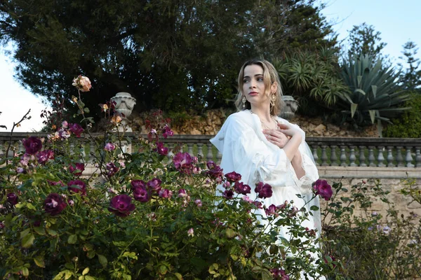 Tarihi beyaz gelinlik ve fantezi pelerin giyen güzel sarışın mankenin portresi. Peri masalı şatosu bahçesinin romantik gül bahçesinde gezinen ruhani figür.