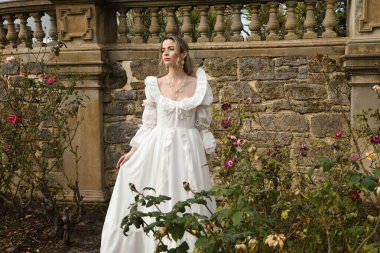 Tarihi fantezi beyaz gelinlik giyen güzel sarışın mankenin portresi. Peri masalı şatosu bahçesinin romantik gül bahçesinde gezinen ruhani figür.