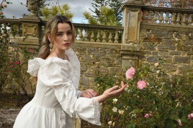 Güzel sarışın mankenin portresi tarihi fantezi beyaz gelinlik giymiş, elleri güllere uzanan, peri masalı kale bahçesindeki gül bahçesi arka planında