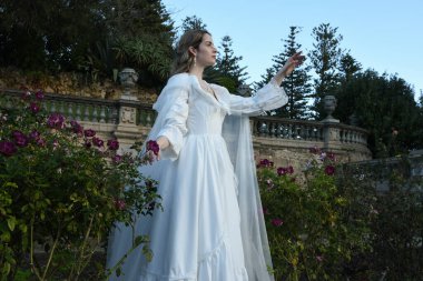 Tarihi beyaz gelinlik ve fantezi pelerin giyen güzel sarışın mankenin portresi. Peri masalı şatosu bahçesinin romantik gül bahçesinde gezinen ruhani figür.