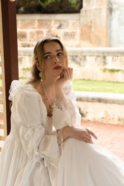 Beyaz fantezi gelinlik giyen sarışın modelin portresi. Tarihsel bir şatonun penceresinden bakan tuhaf bir poz. 