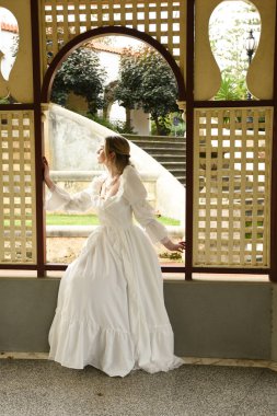 Beyaz fantezi gelinlik giyen sarışın bir kadın figürü. Tarihsel bir şatonun penceresinden bakan tuhaf bir poz. 