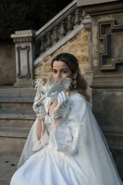 Tarihi beyaz gelinlik, fantezi pelerin ve kelebek maskesi giyen güzel sarışın mankenin portresi. Romantik bir kalenin arka planında gezinen ruhani bir figür.