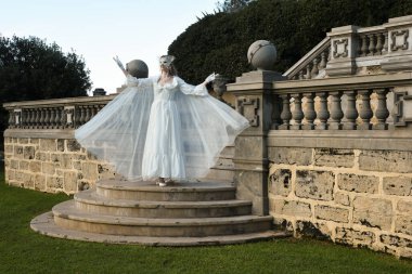 Fantezi beyaz balo elbisesi ve kelebek maskesi giyen güzel sarışın mankenin portresi. Romantik figür tarihi masal kalesinde poz veriyor. 