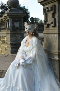 Tarihi beyaz gelinlik, fantezi pelerin ve kelebek maskesi giyen güzel sarışın mankenin portresi. Romantik bir kalenin arka planında gezinen ruhani bir figür.