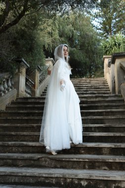 Tarihi beyaz gelinlik ve fantezi pelerin giyen güzel sarışın mankenin portresi. Peri masalı şatosunun tarihi merdivenlerinde yürüyen ruhani bir figür..