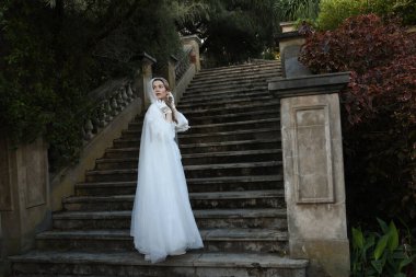 Tarihi beyaz gelinlik ve fantezi pelerin giyen güzel sarışın mankenin portresi. Peri masalı şatosunun tarihi merdivenlerinde yürüyen ruhani bir figür..