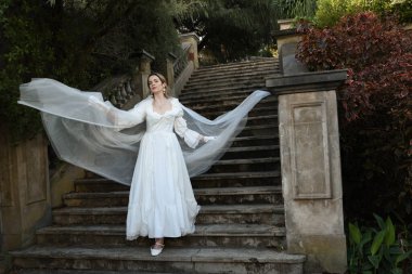 Tarihi beyaz gelinlik ve fantezi pelerin giyen güzel sarışın mankenin portresi. Peri masalı şatosunun tarihi merdivenlerinde yürüyen ruhani bir figür..