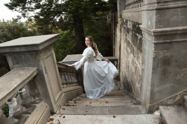 Sarışın güzel kadın modelin tam boy portresi. Tarihi beyaz gotik gelinlik giyiyor. Taş balkonlu romantik masal kalesinin merdivenlerinden iniyor.