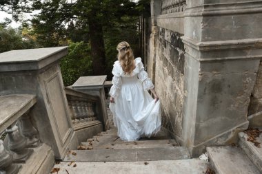 Sarışın güzel kadın modelin tam boy portresi. Tarihi beyaz gotik gelinlik giyiyor. Taş balkonlu romantik masal kalesinin merdivenlerinden iniyor.