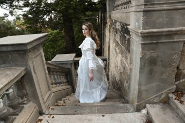 Sarışın güzel kadın modelin tam boy portresi. Tarihi beyaz gotik gelinlik giyiyor. Taş balkonlu romantik masal kalesinin merdivenlerinden iniyor.