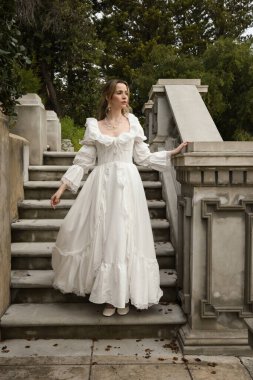 Beyaz gotik gelinlik giyen güzel sarışın mankenin tam boy portresi. Taş balkonlu romantik masal kalesinin merdivenlerinden iniyor.