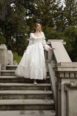 Beyaz gotik gelinlik giyen güzel sarışın mankenin tam boy portresi. Taş balkonlu romantik masal kalesinin merdivenlerinden iniyor.