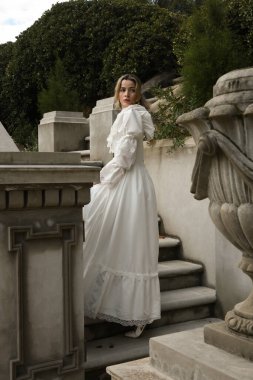 Sarışın güzel kadın modelin tam boy portresi. Tarihi beyaz gotik gelinlik giyiyor. Taş balkonlu romantik masal kalesinin merdivenlerinden iniyor.