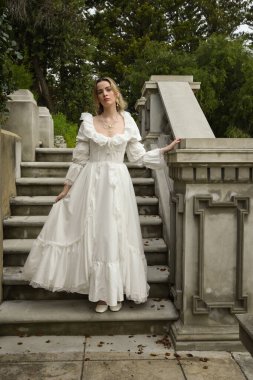 Beyaz gotik gelinlik giyen güzel sarışın mankenin tam boy portresi. Taş balkonlu romantik masal kalesinin merdivenlerinden iniyor.