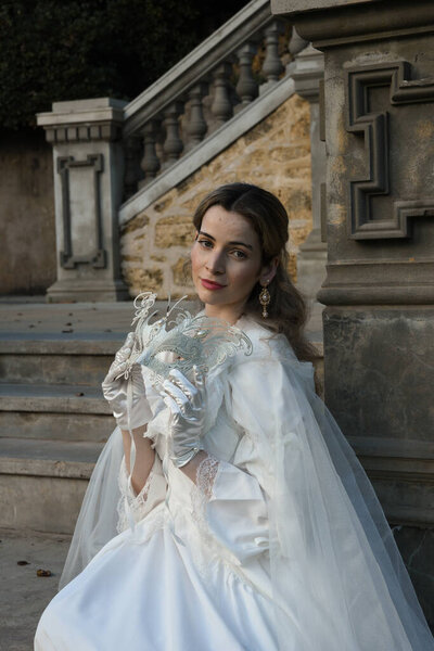 портрет красивой молодой блондинки в историческом белом свадебном платье, фэнтези-плащ и маска-маскарад бабочка. блуждающая эфирная фигура на фоне романтического сказочного сада замка