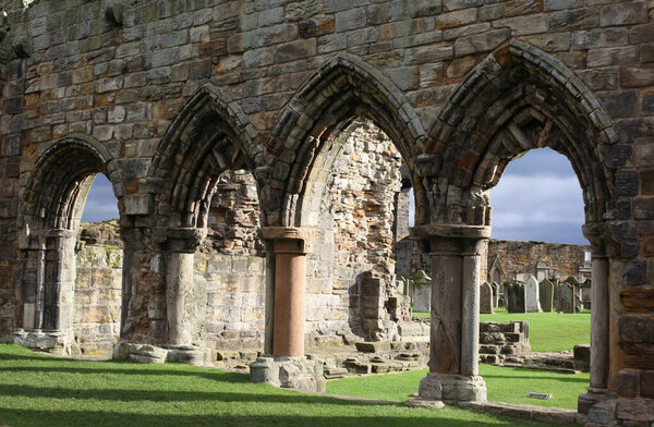 Atmospheric landscape of St Andrews Abbey in Scotland, crumbling gothic architecture surrounded by a haunting graveyard beneath overcast coastal skies.