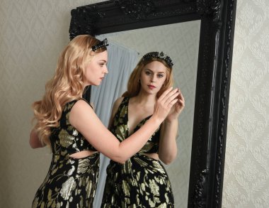 Şık siyah elbisesi olan, fantezi tacıyla sarışın bir kadının portresi. Süslü gotik aynaya bakıyor. Eskimiş desenli duvar kâğıdı arka planı..