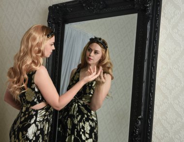 Şık siyah elbisesi olan, fantezi tacıyla sarışın bir kadının portresi. Süslü gotik aynaya bakıyor. Eskimiş desenli duvar kâğıdı arka planı..