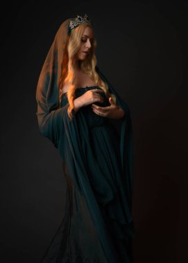 Yeşil balo elbisesi giyen güzel sarışın kadın modelin portresi, peçeli peri masalı tanrıçası kostümü ve kraliyet fantezi tacı. Stüdyonun arka planında izole bir şekilde poz veriyor..
