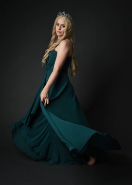 Kraliyet yeşil balo elbisesi giyen sarışın kadın modelin tam boy portresi, fantezi tacı. Durmak, uzaklaşmak, arka plan, karanlık stüdyo arka planında izole bir figür..