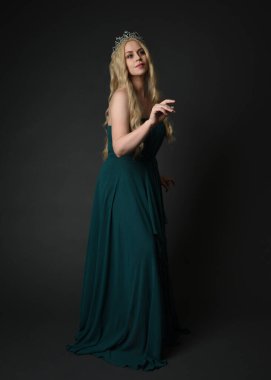 Kraliyet yeşil balo elbisesi giyen sarışın kadın modelin tam boy portresi, fantezi tacı. Durma, yürüme duruşu, ön görüş, karanlık stüdyo arka planında izole bir figür..