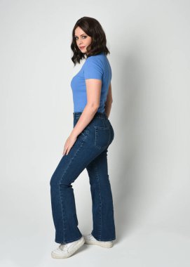 Modern mavi gömlek ve kot pantolon ve spor ayakkabı giyen güzel esmer kadın model. Beyaz stüdyo arka planında, izole edilmiş bir figür olarak geriye doğru yürüyordu..