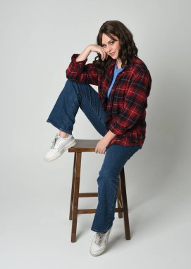 Modern ekose gömlek, kot pantolon ve spor ayakkabı giyen esmer, güzel bir kadın model. İskemlede oturup, beyaz stüdyo arka planında poz veren rahat bir figür.