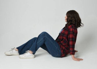 Modern ekose gömlek, kot pantolon ve spor ayakkabı giyen esmer, güzel bir kadın model. Yan profilde oturan, beyaz stüdyo arka planında poz veren rahatlamış yalnız bir figür..