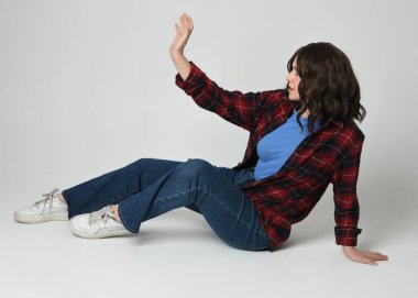 Full length portrait pretty young brunette female model wearing casual modern plaid  flannel shirt and jeans, sneakers. Sitting in side profile, relaxed Isolated figure posing on white studio background.