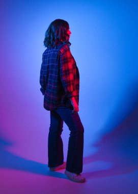 Esmer kadın modelin tam boy modeli gündelik modern giysiler giyiyor, kırmızı ekose gömlek ve kot pantolon giyiyor, arka görüş siluetini aşıyor. Stüdyo arka planında renkli neon mavi ışıklandırma.