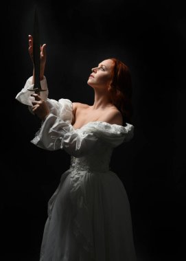 Güzel bir kadın modelin yakın portresi, kızıl saçlı, antika beyaz gelinlik giymiş, hayalet gelin cadılar bayramı karakteri. Karanlık stüdyo arka planında siluet aydınlatma, eller uzanarak ışığa doğru bakıyor.