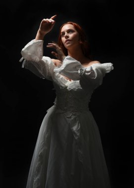 Güzel bir kadın modelin yakın portresi, kızıl saçlı, antika beyaz gelinlik giymiş, hayalet gelin cadılar bayramı karakteri. Karanlık stüdyo arka planında siluet aydınlatma, eller uzanarak ışığa doğru bakıyor.
