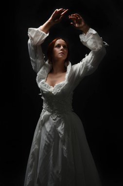 Güzel bir kadın modelin yakın portresi, kızıl saçlı, antika beyaz gelinlik giymiş, hayalet gelin cadılar bayramı karakteri. Karanlık stüdyo arka planında siluet aydınlatma, eller uzanarak ışığa doğru bakıyor.