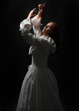 Güzel bir kadın modelin yakın portresi, kızıl saçlı, antika beyaz gelinlik giymiş, hayalet gelin cadılar bayramı karakteri. Karanlık stüdyo arka planında siluet aydınlatma, eller uzanarak ışığa doğru bakıyor.