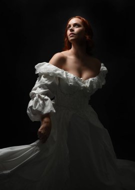 Güzel bir kadın modelin yakın portresi, kızıl saçlı, antika beyaz gelinlik giymiş, hayalet gelin cadılar bayramı karakteri. Karanlık stüdyo arka planında siluet aydınlatma, eller uzanarak ışığa doğru bakıyor.