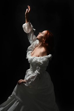 Güzel bir kadın modelin yakın portresi, kızıl saçlı, antika beyaz gelinlik giymiş, hayalet gelin cadılar bayramı karakteri. Karanlık stüdyo arka planında siluet aydınlatma, eller uzanarak ışığa doğru bakıyor.