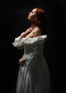 Güzel bir kadın modelin yakın portresi, kızıl saçlı, antika beyaz gelinlik giymiş, hayalet gelin cadılar bayramı karakteri. Karanlık stüdyo arka planında siluet aydınlatma, eller uzanarak ışığa doğru bakıyor.