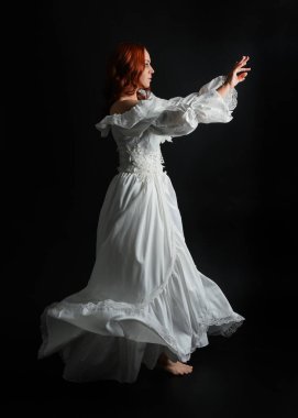 Tam boy portre güzel kadın model antika beyaz hayalet gelinlik giyiyor. Dalgalı kumaş elbiseyle dans pozu. Karanlık stüdyo arka planında izole bir figür.